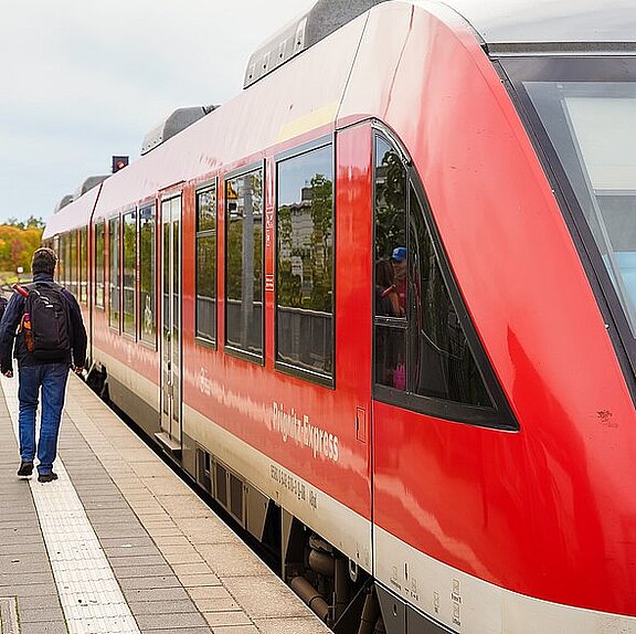 Ein Mann mit einem Rucksack geht entlang eines Bahnsteigs. Rechts steht ein roter Regionalzug, bereit zur Abfahrt. Die Umgebung ist grün und es ist ein bewölkter Tag.