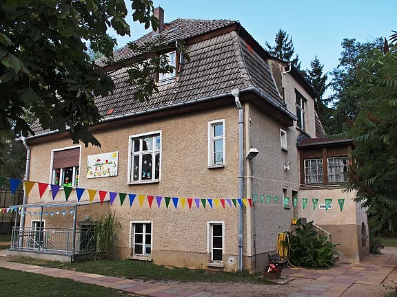 Zweistöckiges, älteres Haus, offenbar als Kindergarten genutzt. An der Fassade ein kindlich gestaltetes Schild; quer über den Hof hängen bunte Wimpelketten. Sommerliche Außenansicht mit etwas Grün und Bäumen - vergrößerte Ansicht in Bildergalerie
öffnen
