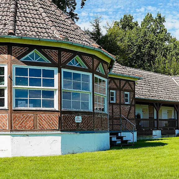 Historisches Fachwerkhaus mit großem, verglastem Pavillon und grauem Ziegeldach. Seitlicher Eingang mit Treppe zu einer überdachten Veranda. Davor gepflegter Rasen, dahinter Bäume bei sonnigem Wetter.