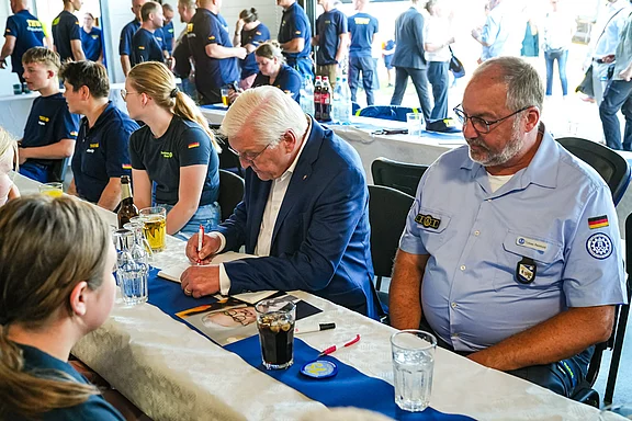 Eine Gruppe von Menschen sitzt an einer Tafel. Ein älterer Mann mit weißen Haaren signiert ein Dokument. Neben ihm sitzt ein weiterer Mann in blauem Hemd. Die Atmosphäre wirkt informell und gesellig. Einige Personen im Hintergrund unterhalten sich.