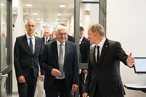 Drei Männer stehen in einem hellen Flur. Der Mann in der Mitte trägt einen Anzug und lächelt, während die anderen beiden ihn anblicken. Der Mann rechts gestikuliert, als ob er etwas erklärt. Im Hintergrund sind weitere Personen und Büromöbel sichtbar.