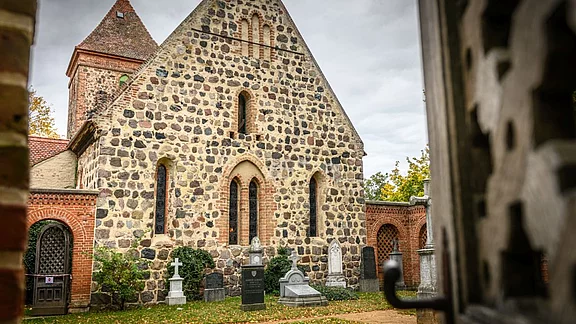 Eine steinerne Kirche mit einem hohen, spitz zulaufenden Dach ist im Hintergrund sichtbar. Vor der Kirche befinden sich mehrere Grabsteine und Bäume. Der Himmel ist bewölkt, was eine ruhige Atmosphäre schafft. Die Eingangstür ist leicht geöffnet.