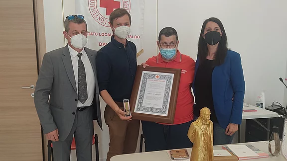 Vier Personen stehen an einem Tisch, der mit der Flagge des Roten Kreuzes dekoriert ist. Sie halten eine Urkunde und posieren zusammen. Im Hintergrund ist ein Banner des Roten Kreuzes sichtbar. Auf dem Tisch stehen verschiedene Gegenstände, darunter eine goldene Statue.