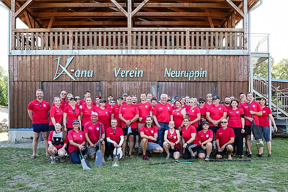 Eine Gruppe von etwa 50 Personen steht vor dem Kanu Verein Neuruppin. Alle tragen rote T-Shirts. Im Hintergrund ist ein Holzgebäude zu sehen. Die Stimmung wirkt freundlich und gemeinschaftlich, als ob die Gruppe für ein Gruppenfoto posiert.