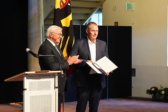 Zwei Männer stehen auf einer Bühne. Der ältere Mann applaudiert, während der jüngere Mann ein Zertifikat in den Händen hält. Im Hintergrund ist eine Fahne mit einem schwarz-gelben Design sichtbar.