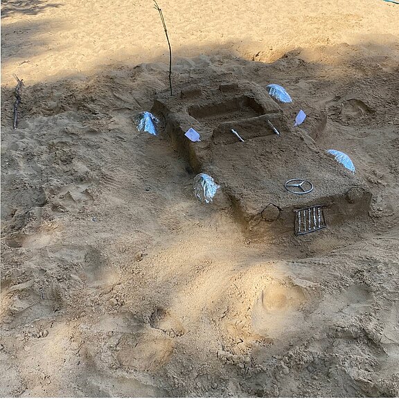 Auf einem Sandstrand steht eine detailreiche Sandskulptur in Form eines Autos. Auf der Motorhaube ist ein Mercedes-Stern gestaltet; Muscheln markieren Scheinwerfer und Kotflügel, ein Stock ragt als Antenne heraus.