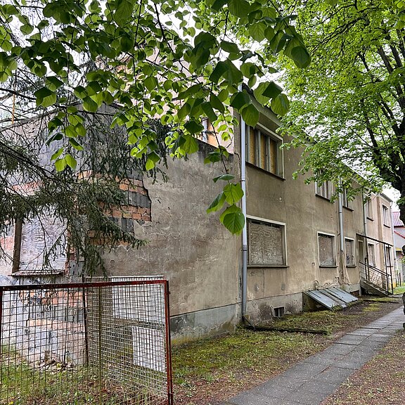 Verfallenes, mehrstöckiges Wohnhaus mit abgeblättertem Putz und verbarrikadierten Fenstern, von Bäumen überschattet. Ein schmaler Gehweg führt entlang der Fassade; Umgebung wirkt verlassen und teilweise überwuchert, vermittelt den Eindruck von Leerstand.