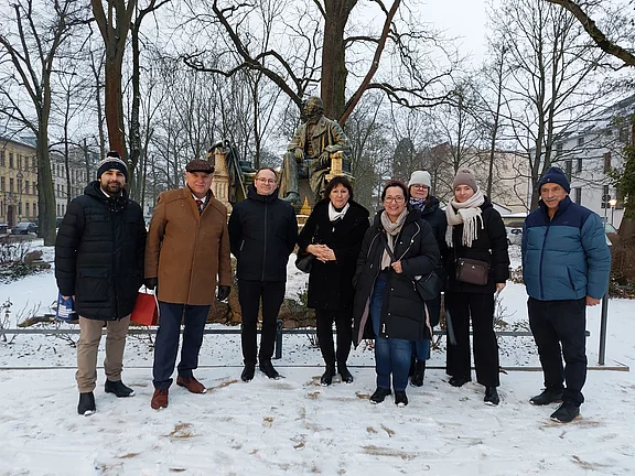 Eine Gruppe von acht Personen steht im Schnee, umgeben von Bäumen. Im Hintergrund ist eine Statue zu sehen. Die Personen lächeln und sind winterlich gekleidet. Es scheint ein Treffen oder eine Veranstaltung im Freien zu sein.