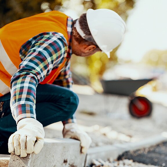 Ein Bauarbeiter mit Schutzhelm, Warnweste und Handschuhen kniet und richtet einen schweren Bordstein an einer Straßenbaustelle aus.
