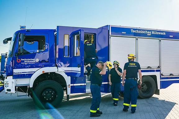 Eine Gruppe von fünf Einsatzkräften des Technischen Hilfswerks (THW) steht um ein blaues THW-Fahrzeug. Einige der Helfer tragen Helme. Einer steigt in das Fahrzeug ein, während ein anderer seine Ausrüstung überprüft.