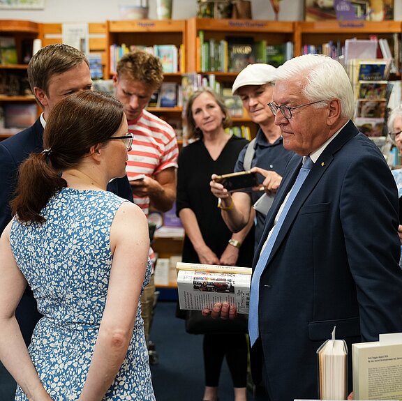 Eine Gruppe von Menschen in einem Bücherladen, die miteinander sprechen. Im Vordergrund steht eine Frau mit Brille und einem blauen Kleid, während ein älterer Mann in Anzug auf sie schaut. Weitere Personen stehen im Hintergrund und beobachten das Geschehen.
