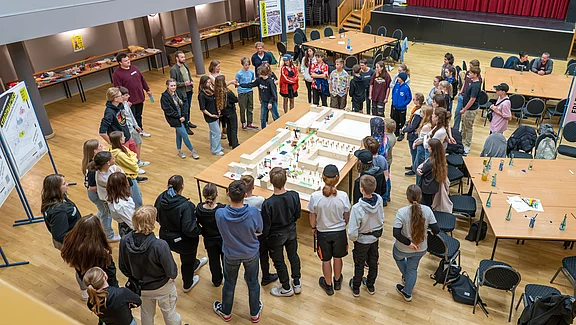 In einem Raum stehen etwa 30 Personen um einen großen Tisch, auf dem ein Modell angeordnet ist. Die Teilnehmer diskutieren und schauen aufmerksam auf das Modell. Die Atmosphäre wirkt interaktiv und gemeinschaftlich, mit verschiedenen Altersgruppen und Geschlechtern vertreten.