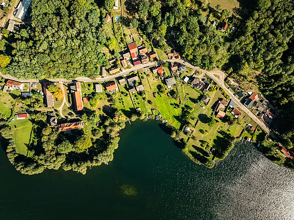 Luftaufnahme eines kleinen, malerischen Dorfes am Ufer eines Sees, umgeben von üppigem Grün. Die Häuser sind mit roten Dächern und Gärten angeordnet. Ein ruhiger Wasserspiegel reflektiert die Umgebung. Der Dorfweg windet sich durch die Landschaft.