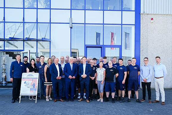 Eine Gruppe von Mitarbeitenden steht vor einem modernen Gebäude. Sie tragen verschiedene Arbeitskleidung und formelle Kleidung. In der Mitte stehen mehrere Männer und Frauen, einige lächelnd, während andere aufmerksam in die Kamera schauen. Im Vordergrund steht ein Plakat.