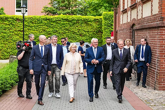 Eine Gruppe von Personen, darunter mehrere Männer in Anzügen und eine Frau in einer hellen Jacke, geht gemeinsam auf einem gepflasterten Weg. Im Hintergrund sind grüne Bäume und eine Gebäudeansicht sichtbar. Einige Personen scheinen Kameras zu tragen.