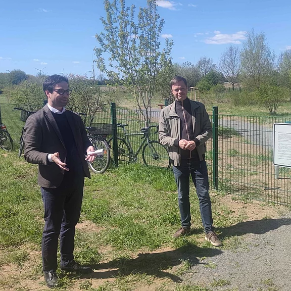 Zwei Männer stehen in einer grünen Parklandschaft neben einem Zaun. Der linke spricht und gestikuliert mit Notizzetteln, der rechte hört zu. Hinter ihnen lehnen Fahrräder. Sonniger Tag.