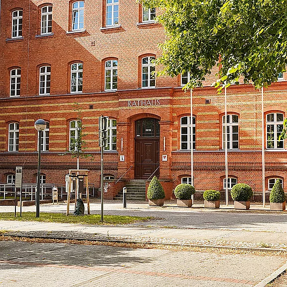 Ein rotes Backsteingebäude mit der Aufschrift "RATHAUS" über dem Eingang. Vor dem Gebäude befinden sich symmetrisch angeordnete Bäume in Pflanzkübeln und ein paar Lampen. Die Fassade zeigt große Fenster und eine einladende Eingangstür.