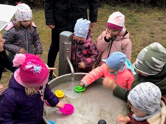 Eine Gruppe von Kindern steht um ein Wasserbecken im Freien. Eine erwachsene Person, die sie betreut, lächelt sie an. Die Kinder spielen mit bunten Bechern im Wasser und tragen warme Kleidung. Der Hintergrund zeigt eine eingezäunte Grünfläche.