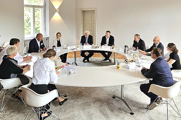 Eine Gruppe von Personen sitzt um einen runden Tisch in einem hellen Raum. Sie sind in formeller Kleidung gekleidet und scheinen an einem Gespräch oder Meeting teilzunehmen. Einige Teilnehmer notieren Informationen, während Getränke und Unterlagen auf dem Tisch verteilt sind.