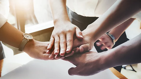 Mehrere Hände liegen übereinander auf einem Tisch. Die Hände stammen von verschiedenen Personen und symbolisieren Zusammenarbeit und Teamgeist. Es gibt eine Uhr an einem Handgelenk. Die Szene vermittelt ein Gefühl von Einheit und Gemeinschaft.