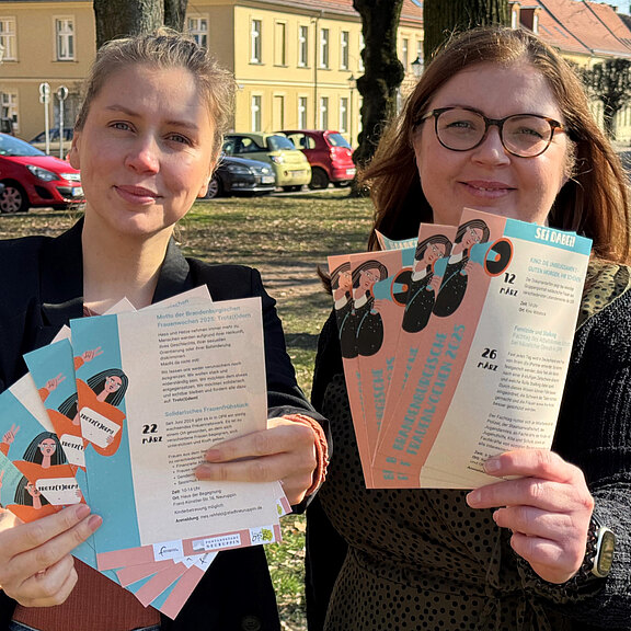 Zwei Frauen stehen im Freien und halten mehrere Informationsbroschüren in den Händen. Die Broschüren sind farbenfroh gestaltet und enthalten Texte zur Präsentation. Im Hintergrund sind Bäume und Fahrzeuge zu sehen.