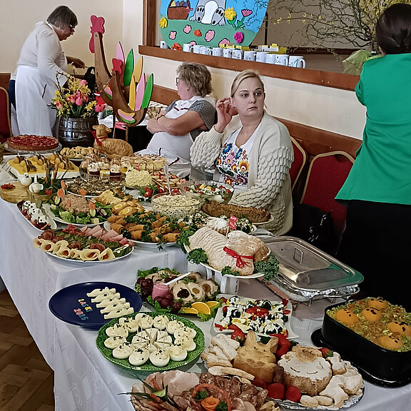 Reich gedecktes Buffet in einem Gemeindesaal: zahlreiche kalte Platten, gefüllte Eier, Salate, Kuchen und Desserts. Mehrere Frauen ordnen die Speisen. Frühlingshafte, teils österliche Dekoration im Hintergrund. Fokus auf Vielfalt und Fülle der Speisen.