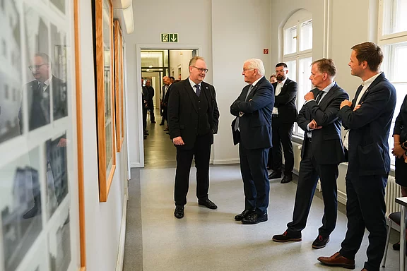 Eine Gruppe von Männern in Anzügen steht in einem schmalen Flur. Sie unterhalten sich angeregt, während sie Bilder an den Wänden betrachten. Im Hintergrund sind weitere Personen zu sehen, die ebenfalls an der Diskussion teilnehmen.