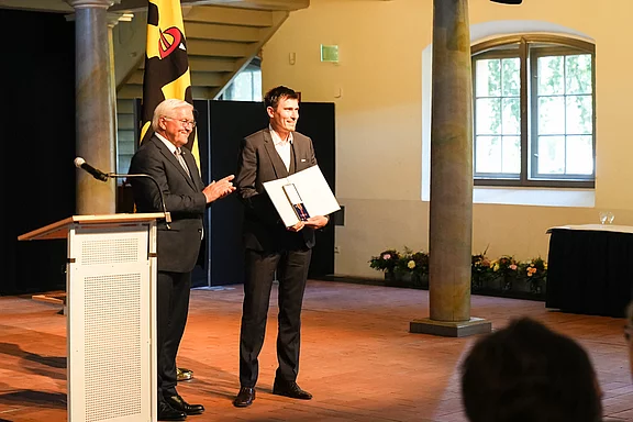 Ein Mann in Anzug steht auf einer Bühne und hält eine Urkunde in der Hand. Neben ihm steht ein älterer Mann, der applaudiert. Im Hintergrund sind Vorhänge und Fenster sichtbar. Die Szene hat einen feierlichen Charakter, vermutlich eine Auszeichnung oder Ehrung.