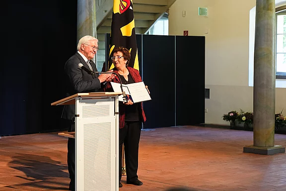 Ein älterer Mann spricht an einem Rednerpult, während eine Frau neben ihm mit einer Urkunde steht. Im Hintergrund ist ein schwarzes Tuch und eine Flagge sichtbar. Beide Personen scheinen an einem feierlichen Anlass teilzunehmen, möglicherweise einer Preisverleihung oder Ehrung.