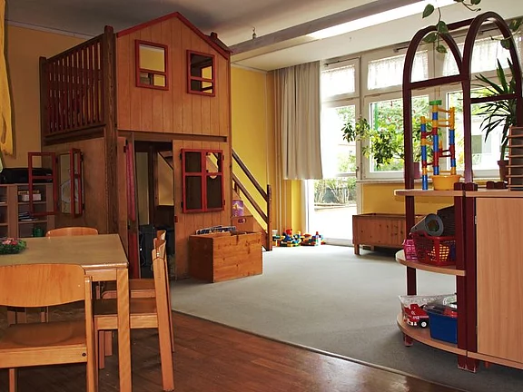 Ein helles Spielzimmer mit einer Holzspielburg auf zwei Ebenen und roten Fenstern. Im Vordergrund stehen ein Tisch und Stühle. In der Ecke sind Spielzeugkisten und ein Regal mit bunten Bausteinen. Fenster lassen Tageslicht herein, und einige Pflanzen sind sichtbar.
