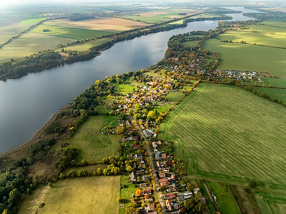 Luftaufnahme eines ländlichen Gebiets mit einem Fluss, der durch die Landschaft fließt. Am Ufer sind mehrere kleine Häuser und ein Dorf zu sehen, umgeben von Feldern und Bäumen. Die Landschaft zeigt verschiedene Grüntöne und einige Herbstfarben.