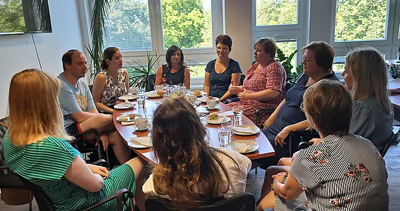 Mehrere Erwachsene sitzen in einem Kreis um einen Tisch und führen eine lebhafte Gesprächsrunde. Auf dem Tisch stehen Tassen, Teller und kleine Speisen. Die Szene wirkt wie ein informelles Treffen oder Workshop in einem hellen Raum.