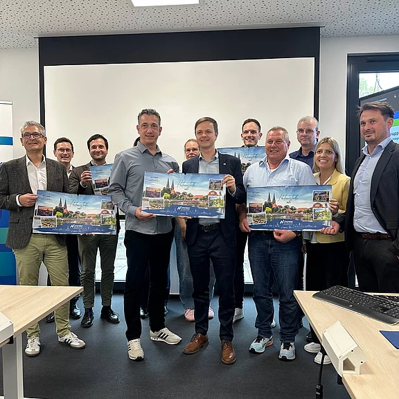 Etwa zwölf Erwachsene posieren in einem Konferenzraum; mehrere halten breite Plakate mit Stadtansichten. Die Gruppe wirkt feierlich, als ginge es um eine Präsentation oder Übergabe.