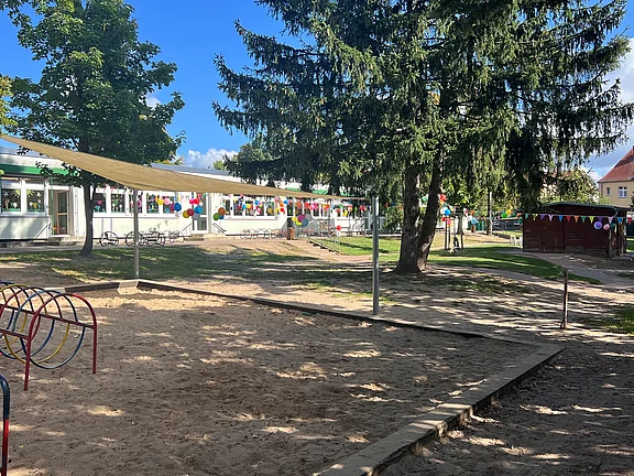Der Blick auf einen Spielplatz mit einem Sandkasten und Klettergerüst im Vordergrund. Im Hintergrund steht ein mehrstöckiges Gebäude, umgeben von Bäumen und bunten Fähnchen, die festlich dekoriert sind. Der Himmel ist blau mit einigen Wolken.