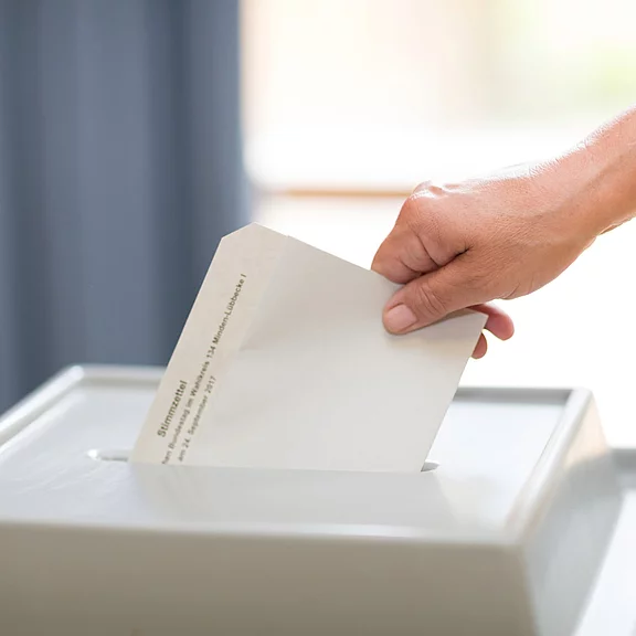 Nahaufnahme: Eine Hand schiebt einen Stimmzettel in den Schlitz einer Wahlurne – Symbol für eine laufende Abstimmung.