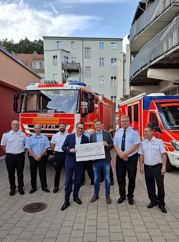 Gruppe von sechs Feuerwehrmännern und zwei Zivilpersonen steht vor zwei roten Feuerwehrfahrzeugen. In der Mitte halten zwei Männer einen großen Spendenscheck, während alle in die Kamera blicken. Die Szene zeigt eine offizielle Spendenübergabe an die Feuerwehr im Freien.