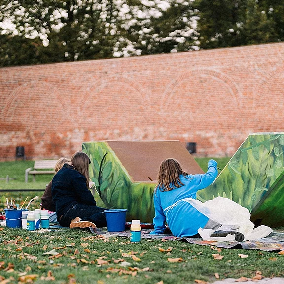 Mehrere Kinder malen im Freien eine große grüne Skulptur. Sie sitzen auf dem Rasen, tragen Malkittel und arbeiten mit Pinseln und Farbtöpfen. Die Szene zeigt ein gemeinsames Kunstprojekt im Außenbereich.