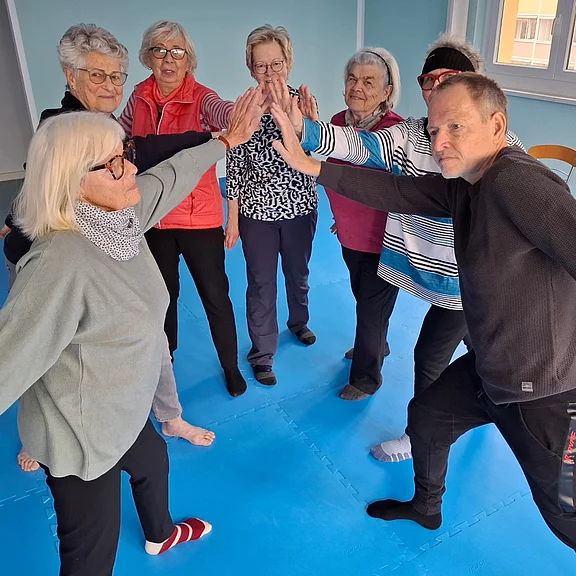 Gruppe älterer Teilnehmerinnen und ein Mann stehen im Kreis in einem Übungsraum. Sie strecken die Arme zur Mitte und geben sich ein gemeinsames High-Five. Die Szene vermittelt Bewegung, Zusammenarbeit und Freude.