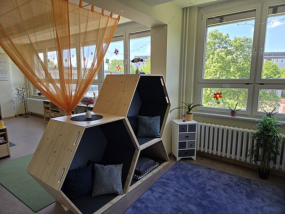 Heller Raum mit zwei übereinander angeordneten, sechseckigen Holznischen mit dunkler Polsterung und Kissen – eine gemütliche Lese- oder Ruheecke. Daneben große Fenster, etwas Grünpflanzen und ein kleines Schränkchen. Insgesamt wirkt der Bereich freundlich und einladend.