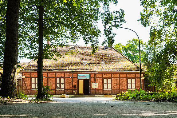Ein historisches, rot-brick Gebäude mit einem Satteldach, umgeben von grünen Bäumen. Die Fassade zeigt Fenster mit weißen Rahmen und eine Eingangstür in der Mitte. Ein Schild mit der Aufschrift „SPEEDOS“ ist deutlich sichtbar über dem Eingang.