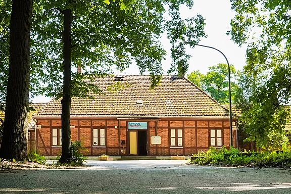 Ein historisches, rot-brick Gebäude mit einem Satteldach, umgeben von grünen Bäumen. Die Fassade zeigt Fenster mit weißen Rahmen und eine Eingangstür in der Mitte. Ein Schild mit der Aufschrift „SPEEDOS“ ist deutlich sichtbar über dem Eingang.
