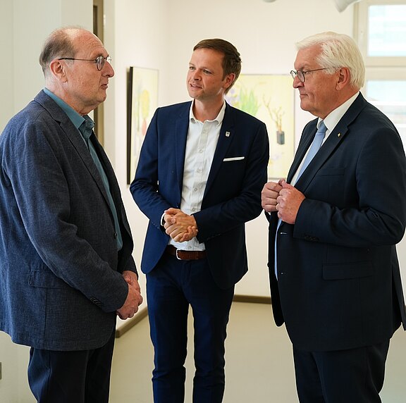 Drei Männer stehen in einem hellen Raum und unterhalten sich. Der Mann links trägt eine Brille und einen blauen Blazer, der in der Mitte hat einen dunklen Anzug und das Hemd offen, während der Mann rechts in einem dunklen Anzug mit einer Krawatte gekleidet ist. Er wirkt freundlich und interessiert.