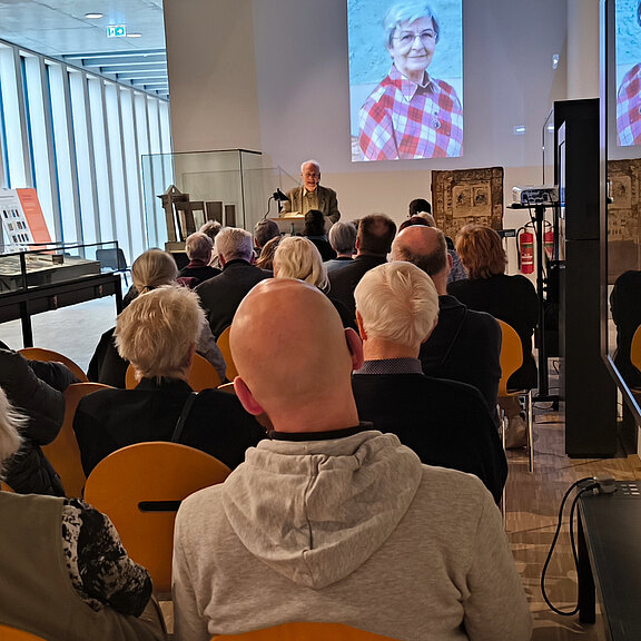 Vortrag in einem Saal: Zuhörer sitzen in Reihen, vorne spricht ein Mann am Rednerpult. Auf der Leinwand hinter ihm erscheint ein großes Porträt einer älteren Frau im karierten Hemd.