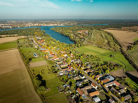 Luftaufnahme einer ländlichen Gegend, die ein Flussbett, ein Dorf mit bunten Häusern und grünen Feldern zeigt. Im Hintergrund erstrecken sich Wälder und Felder bis zum Horizont, unter einem klaren blauen Himmel.