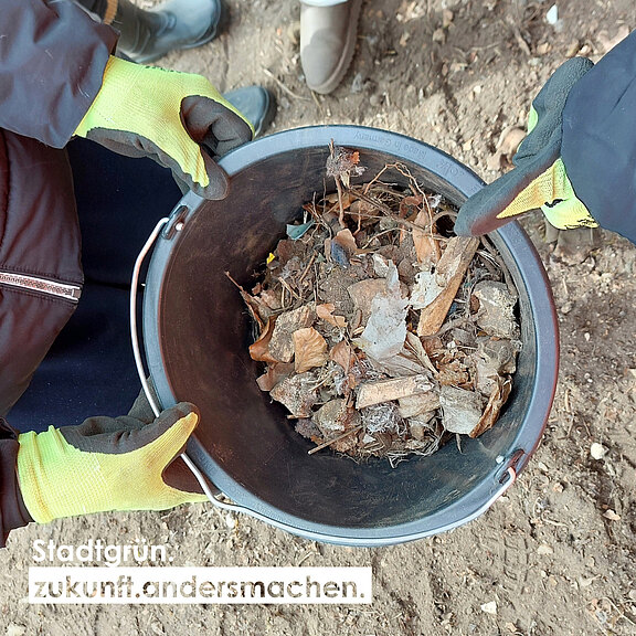 Ein Mittelsmann hält einen Eimer mit trockenem Laub und kleinen Ästen. Die Hände sind mit Arbeitshandschuhen geschützt. Der Hintergrund zeigt den Boden, während eine andere Person leicht im Bild zu sehen ist. Der Schriftzug "Stadtgrün zukunft.andersmachen." ist unten sichtbar.