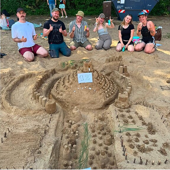 Sechs Erwachsene sitzen am Strand hinter einer großen, detailreichen Sandburg mit vielen Türmen, Gräben und Wegen. Sie lächeln und zeigen Daumen hoch, als würden sie stolz ihr Bauwerk präsentieren.