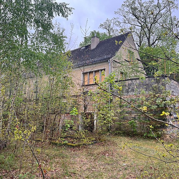 Verwildertes, verlassen wirkendes Wohnhaus am Waldrand. Teilweise verdeckt von dichtem Gestrüpp und jungen Bäumen, mit grauem Ziegeldach und blasser Fassade. Eine hohe, bemooste Steinmauer und rankende Pflanzen umgeben das Gebäude. Bewölkter Himmel verstärkt den vernachlässigten, stillen Eindruck.