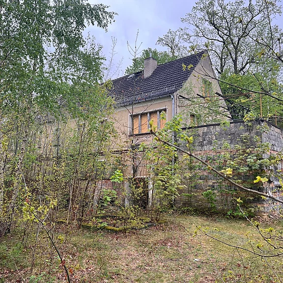 Verwildertes, verlassen wirkendes Wohnhaus am Waldrand. Teilweise verdeckt von dichtem Gestrüpp und jungen Bäumen, mit grauem Ziegeldach und blasser Fassade. Eine hohe, bemooste Steinmauer und rankende Pflanzen umgeben das Gebäude. Bewölkter Himmel verstärkt den vernachlässigten, stillen Eindruck.
