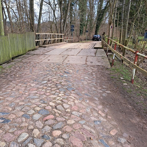 Eine schmale, gepflasterte Straße führt über eine kleine Brücke. Auf der rechten Seite steht ein schwarzes Auto. Am Wegesrand sind ein Zaun und Verkehrszeichen zu sehen. Umgebende Bäume deuten auf eine ländliche Umgebung hin.