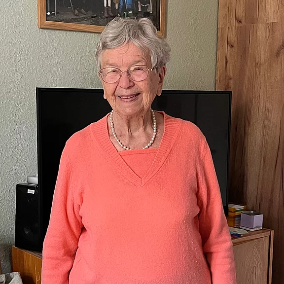 Ältere Frau mit grauen Haaren und Brille lächelt in einer Wohnung. Sie trägt einen korallenfarbenen Pullover und eine Perlenkette.